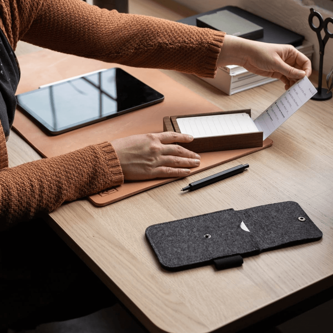 Person placing task list card into walnut card holder next to felt carrying case on leather desk pad.